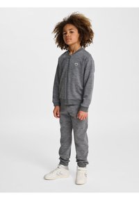 Grå zipjacka med matchande sweatpants; mjukt tyg, ribbade muddar och en broderad logotyp på jackans bröst; vita sneakers.