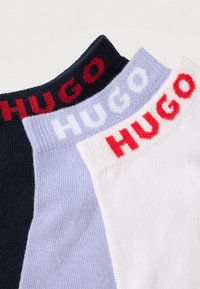 Tři páry ponožek s žebrovanými lemy; barvy zahrnují tmavě modrou, světle modrou a bílou. Každý pár má značku "HUGO" v kontrastních barvách.
