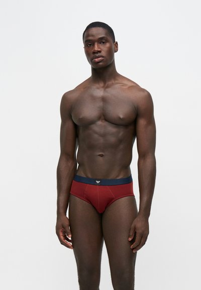 Homme musclé debout devant un fond uni portant un slip rouge foncé avec une ceinture bleu marine.