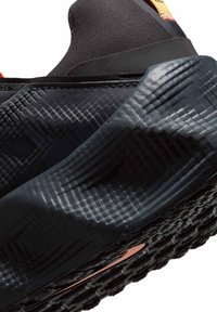 Gros plan de la semelle texturée et de la zone du talon d'une basket noire, montrant des motifs géométriques complexes et un petit détail orange en dessous.