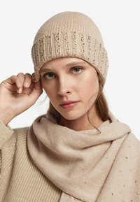 Beżowa dzianinowa czapka z prążkowaną fakturą i cekinowymi akcentami. Modelka nosi pasujący beżowy szalik, tworząc spójny, delikatny wygląd.