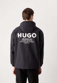 Mustahoodiga must jakk elastsete mansettidega, millel on seljal suur valge "HUGO" bränding ja täiendav tekst, sile tekstuur.