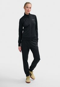 Zwarte sportieve trainingspak van glad materiaal met een ritsjack en bijpassende broek, met een strakke pasvorm en een minimaal logo.