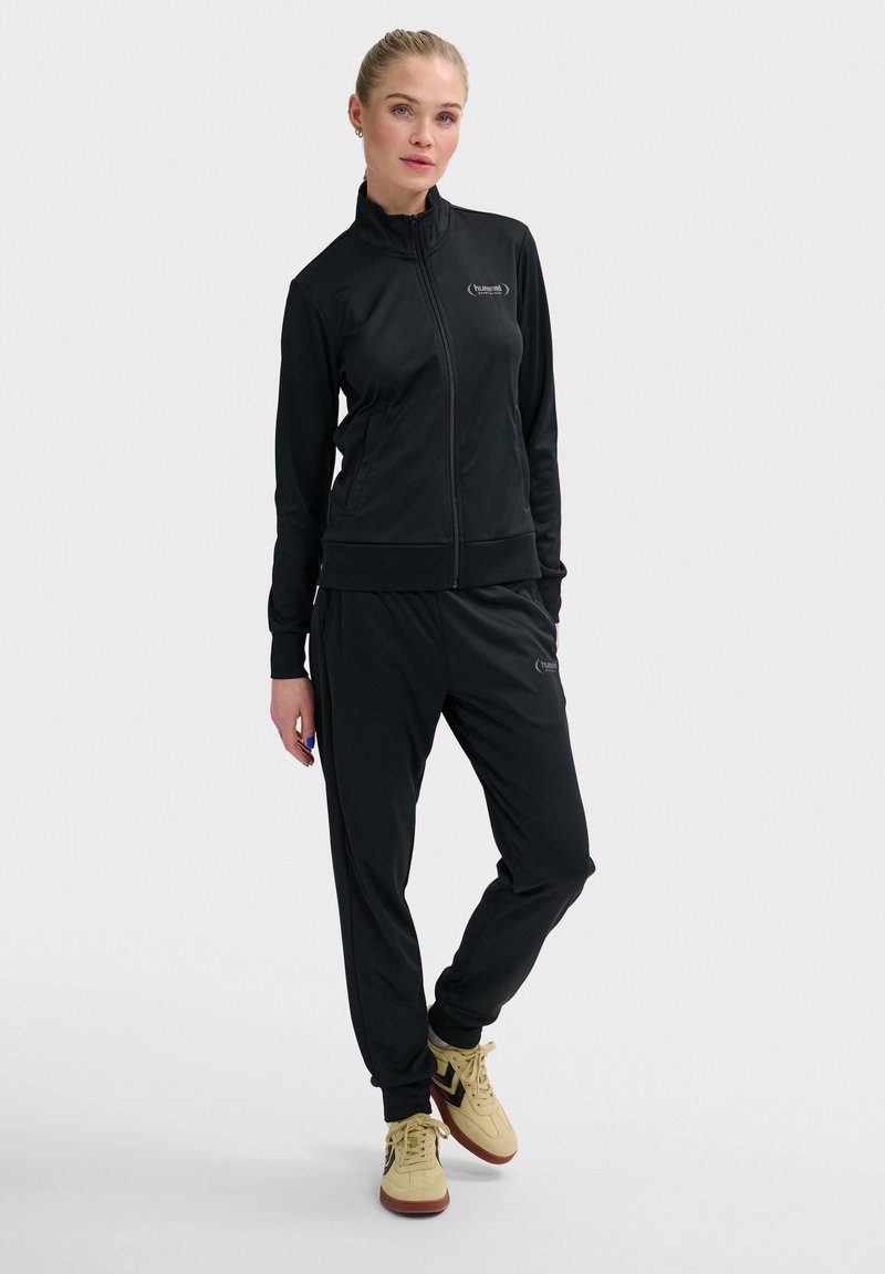 Zwarte sportieve trainingspak van glad materiaal met een ritsjack en bijpassende broek, met een strakke pasvorm en een minimaal logo.