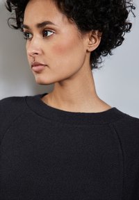 Pull en maille noir avec un col rond et des manches raglan, fabriqué dans un tissu texturé et à coupe décontractée, mettant l'accent sur le confort.