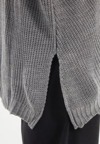 Pull gris tricoté avec une maille lâche, ourlet côtelé et fentes latérales pour faciliter les mouvements. Tissu texturé offrant une apparence décontractée.