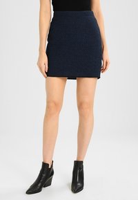 Minifalda azul marino con un patrón texturizado, de cintura alta y corte recto. Combinada con botas negras de tobillo. Tejido suave con un ligero brillo.