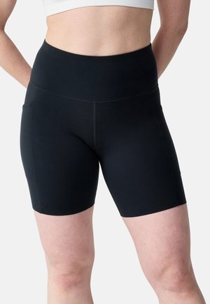 Femme portant un short de sport noir taille haute avec poches latérales et un haut court blanc, debout les mains derrière le dos.