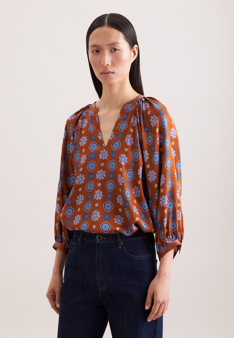 Femme aux cheveux noirs raides, portant un chemisier marron avec des motifs floraux bleus et blancs, et un jean bleu foncé, debout devant un fond uni.