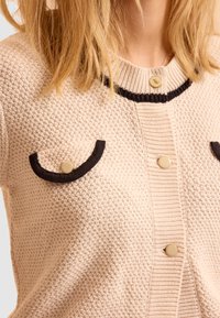 Cardigan lavorato a maglia di un beige chiaro con un motivo testurizzato, caratterizzato da accenti marroni scuri e due tasche frontali. Chiusura a bottone sul colletto.