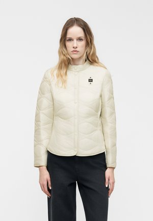 Femme portant une veste matelassée crème avec des boutons pression, debout devant un fond blanc, associée à un pantalon noir.