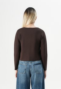 Persoon met lang blond haar, gekleed in een donkerbruin lange mouwen top en blauwe spijkerbroek, gezien van achteren op een witte achtergrond.