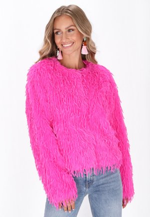 Glijdende vrouw met een helderroze fluffy trui, lichtblauwe jeans en roze franjesoorbellen, kijkt opzij tegen een witte achtergrond.