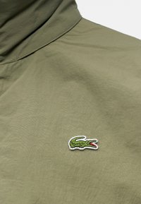 Tissu vert olive avec un petit logo crocodile vert brodé avec une bouche rouge sur la zone de la poitrine d’un vêtement.