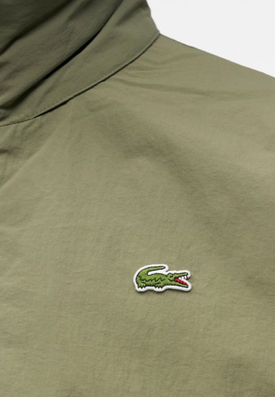 Tissu vert olive avec un petit logo crocodile vert brodé avec une bouche rouge sur la zone de la poitrine d’un vêtement.
