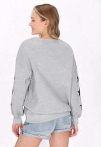 Felpa grigia con maniche lunghe, caratterizzata da motivi a stelle nere lungo le maniche, scollatura rotonda e vestibilità comoda. Abbinata a pantaloni cargo in denim.
