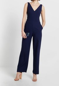 Mörkblå jumpsuit med V-ringning, ärmlös design, vida byxor och sidofickor. Tillbehör inkluderar en liten, färgglad clutch.