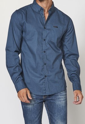 Homme portant une chemise bleue à motifs à manches longues avec des boutons noirs et un jean bleu délavé, debout devant un fond uni.