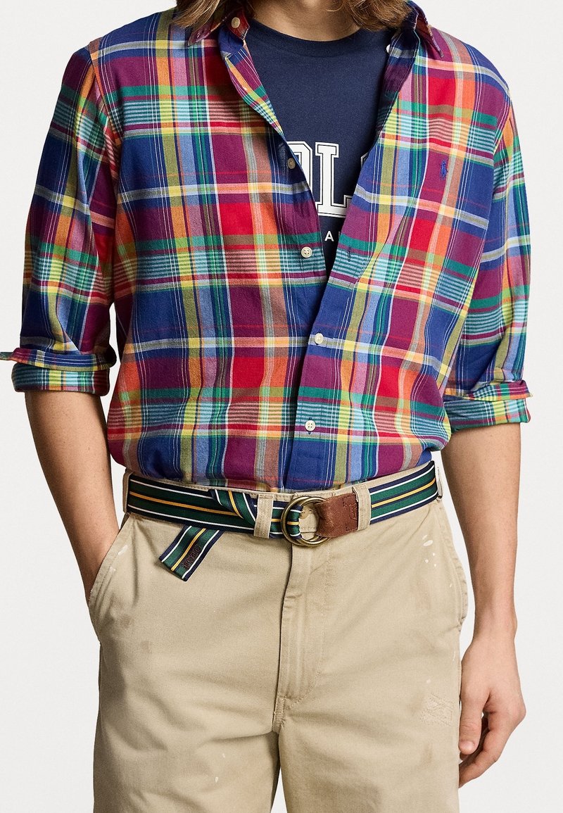 Personne portant une chemise à carreaux multicolores boutonnée sur un t-shirt graphique bleu marine, un pantalon beige et une ceinture en tissu rayé.
