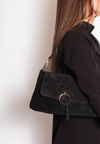 Borsa a tracolla in morbido camoscio nero con design a pattina, caratterizzata da un dettaglio ad anello dorato e da una tracolla removibile. Texture liscia e forma funzionale.