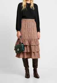Blouse noire à manches longues, jupe rose à fleurs en tiers avec volants, sac à main en cuir croco vert, et bottes marron hautes jusqu'aux genoux.