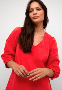 Röd, löst sittande blus med V-ringning, rynkade ballongsärmar och en slät textur. Händerna placerade fram, vilket framhäver plaggets design.