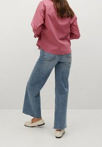 Chemise rose à manches longues, associée à un jean bleu clair à jambes larges. Le jean présente des ourlets effilochés ; les chaussures sont des mocassins blancs avec des semelles noires.