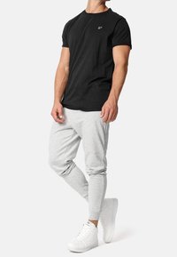 Mand iført sort t-shirt med korte ærmer, lysegrå joggerbukser og hvide høje sneakers, der står med hænderne afslappet.