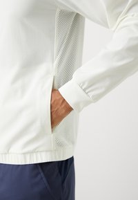 Lett hvid pullover med et tekstureret mesh-panel på siden, ribbede manchetter og sidelommer, parret med mørkeblå bukser.