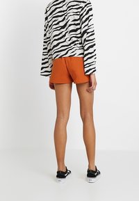 Top a maniche lunghe a strisce zebrate bianco e nero abbinato a pantaloni shorts arancioni. Modella che indossa sneakers nere, mostrando la vista posteriore.