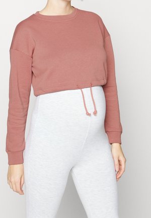 Personne portant un haut court à manches longues couleur rose poussiéreuse et des leggings taille haute gris clair sur un fond uni.