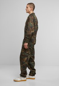 Zöld és barna árnyalatokban készült kamuflázs overáll, cipzáras zsebbel, elasztikus mandzsettákkal és laza szabással. Fehér sportcipővel viselt.