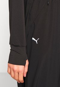 Svart atletisk jacka med långa ärmar, utrustad med en dragkedja och en vit Puma-logotyp på sidan. Tillverkad av mjukt, flexibelt tyg.