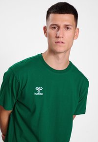 Groene katoen t-shirt met een ronde hals en korte mouwen. Witte geborduurde logo aan de linkerkant van de borst. Gladde textuur.