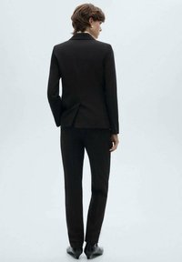 Completo tailored nero con giacca aderente e pantaloni dritti. Presenta una leggera lucentezza, ventaglio singolo sul retro e calzature eleganti.