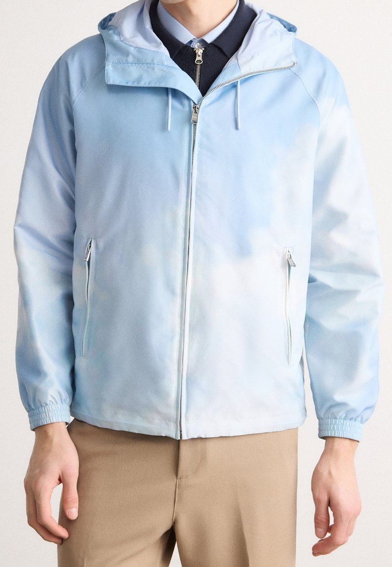 Homme portant une veste à capuche zippée bleu clair avec poches latérales zippées sur une chemise sombre à col et un pantalon beige.