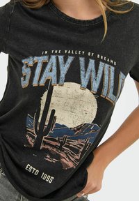 Zwarte graphic t-shirt met de tekst "STAY WILD", met een woestijnlandschap met cactussen en bergen, een verouderd ontwerp en een ronde halslijn.