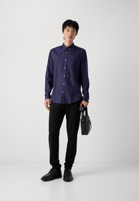 Cămașă cu mâneci lungi, cu nasturi, de culoare albastru închis, cu un model geometric, purtată cu pantaloni negri și pantofi negri, ținând o geantă mică neagră.