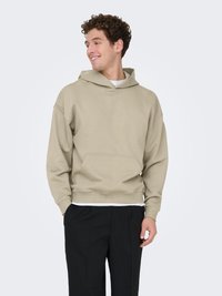 Sweat à capuche beige avec une grande poche frontale, poignets et ceinture côtelés. Tissu à texture lisse, coupe décontractée, porté par-dessus une chemise blanche.