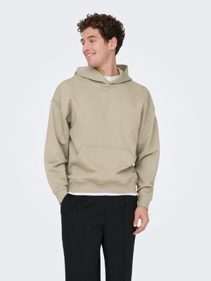 Felpa beige con cappuccio, ampie tasche frontali, polsini e orlo a costine. Tessuto dalla texture liscia, vestibilità rilassata, indossata sopra una maglietta bianca.