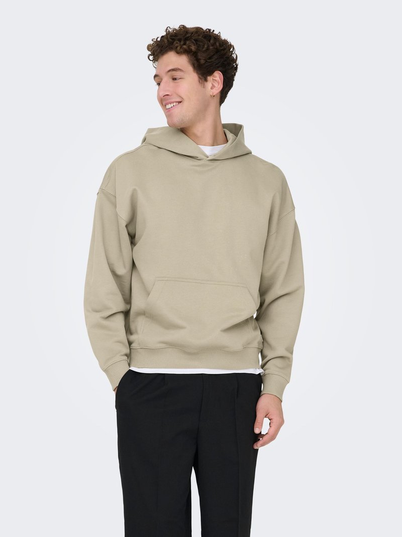 Sweat à capuche beige avec une grande poche frontale, poignets et ceinture côtelés. Tissu à texture lisse, coupe décontractée, porté par-dessus une chemise blanche.