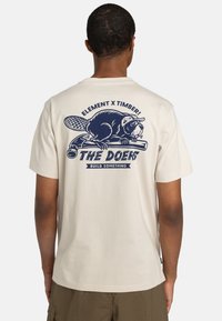 Beige Baumwoll-T-Shirt mit einem navyfarbenen Grafikdruck auf der Rückseite, der einen Biber, Werkzeuge und den Text "DIE MACHER BAUEN ETWAS" zeigt. Kurzärmlig.