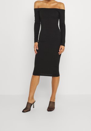 Femme portant une robe midi noire à épaules dénudées et manches longues, ainsi que des chaussures à talons hauts noires en maille à bout ouvert, debout sur un fond uni.