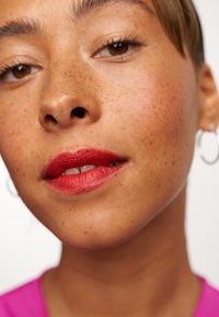 Gros plan sur un visage avec une peau brun clair, des taches de rousseur et des lèvres rouges. La personne porte de petites boucles d'oreilles en argent et un haut rose vif.