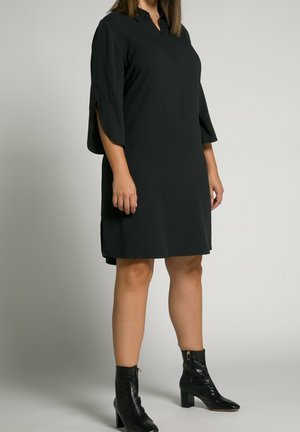 Kvinde iført en knælængde sort kjole med trekvart lange ærmer med slids og sorte ankelstøvler med hæl, stående mod en ensfarvet baggrund.