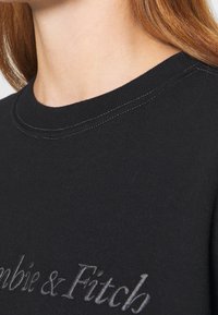 Sudadera negra con cuello redondo, textura suave y costuras en blanco contrastante. Presenta texto bordado en un tono más claro.