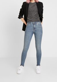 Ljusblå skinny jeans med hög midja, tillsammans med en svart kavaj och en glittrig grå topp. Vita sneakers fulländar outfiten.