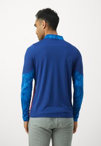 Blauwe sportshirt met lange mouwen, met patronen op de mouwen, textiel met structuur en een hoge kraag. Gecombineerd met grijze sportbroek.