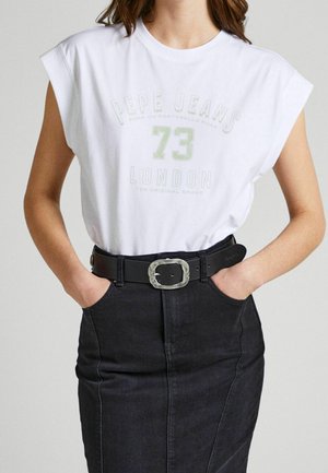 Kobieta nosząca białą bezrękawnikową koszulkę Pepe Jeans 73 London z grafiką, wpuszczoną w czarną jeansową spódnicę z wysokim stanem i czarnym paskiem.