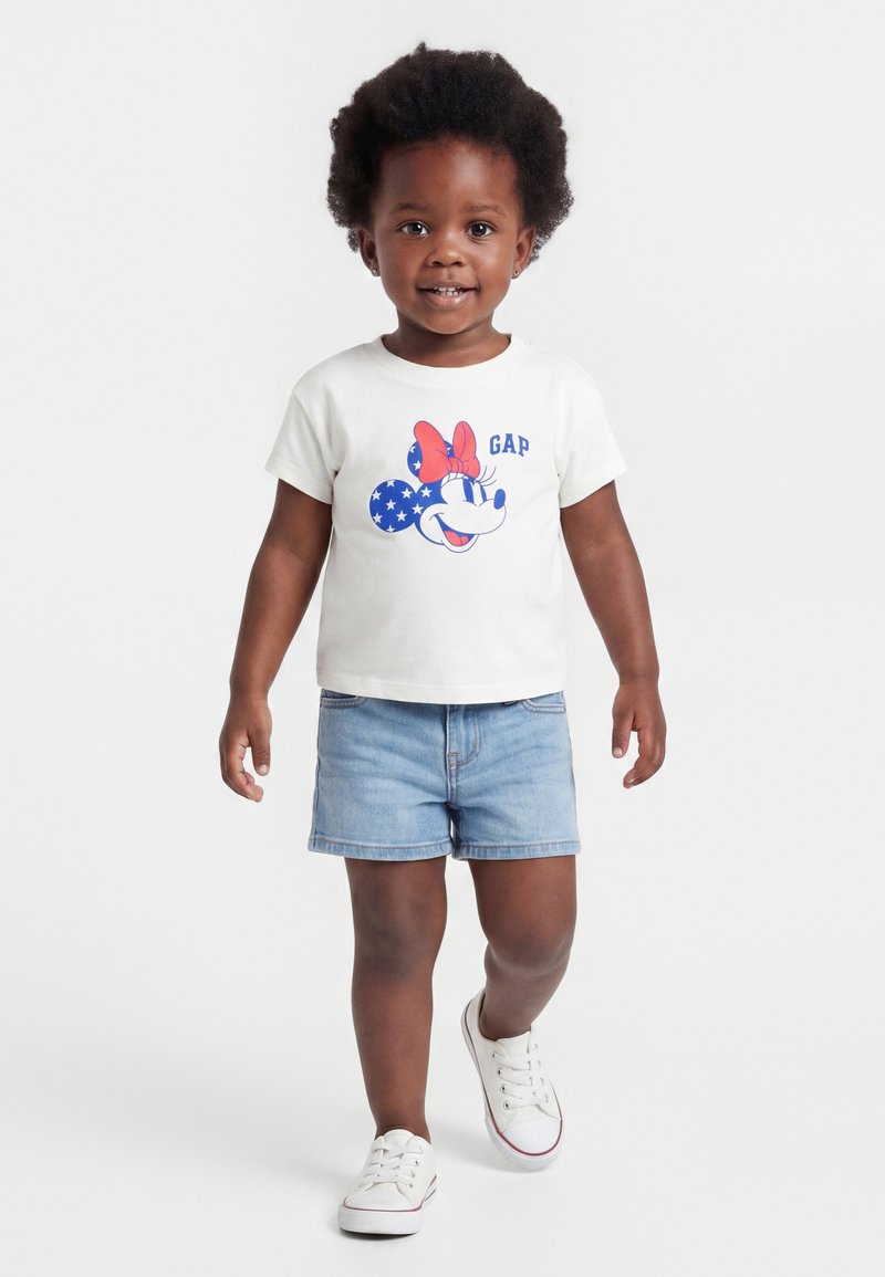 Νήπιο με λευκό t-shirt Minnie Mouse, τζιν σορτς και λευκά αθλητικά παπούτσια περπατά προς τα εμπρός σε απλό φόντο.